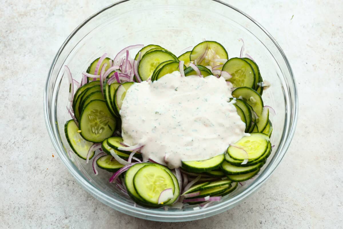 Creamy Cucumber Salad is a delicious and refreshing side dish. All it takes is a handful of fresh simple ingredients and 15 minutes. This is the perfect addition to your next summer barbecue! Dressing poured over cucumbers, ready to combine.