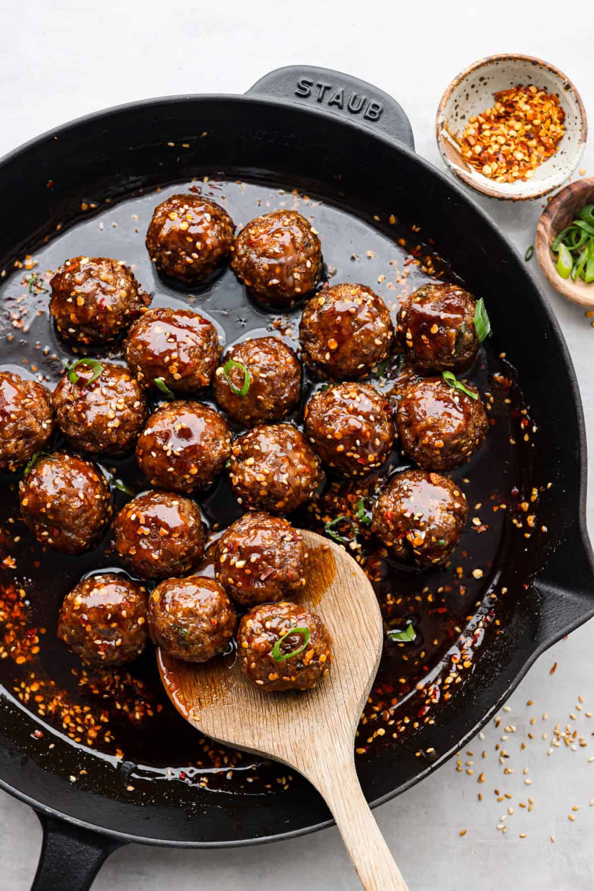Get ready to spice up your dinner with these firecracker meatballs! Juicy beef meatballs are baked to perfection and then tossed in a sweet and spicy buffalo-based sauce, creating a delicious meal that's perfect for any night of the week. overhead shot of firecracker meatballs doused in sauce in a skillet with wooden spoon.