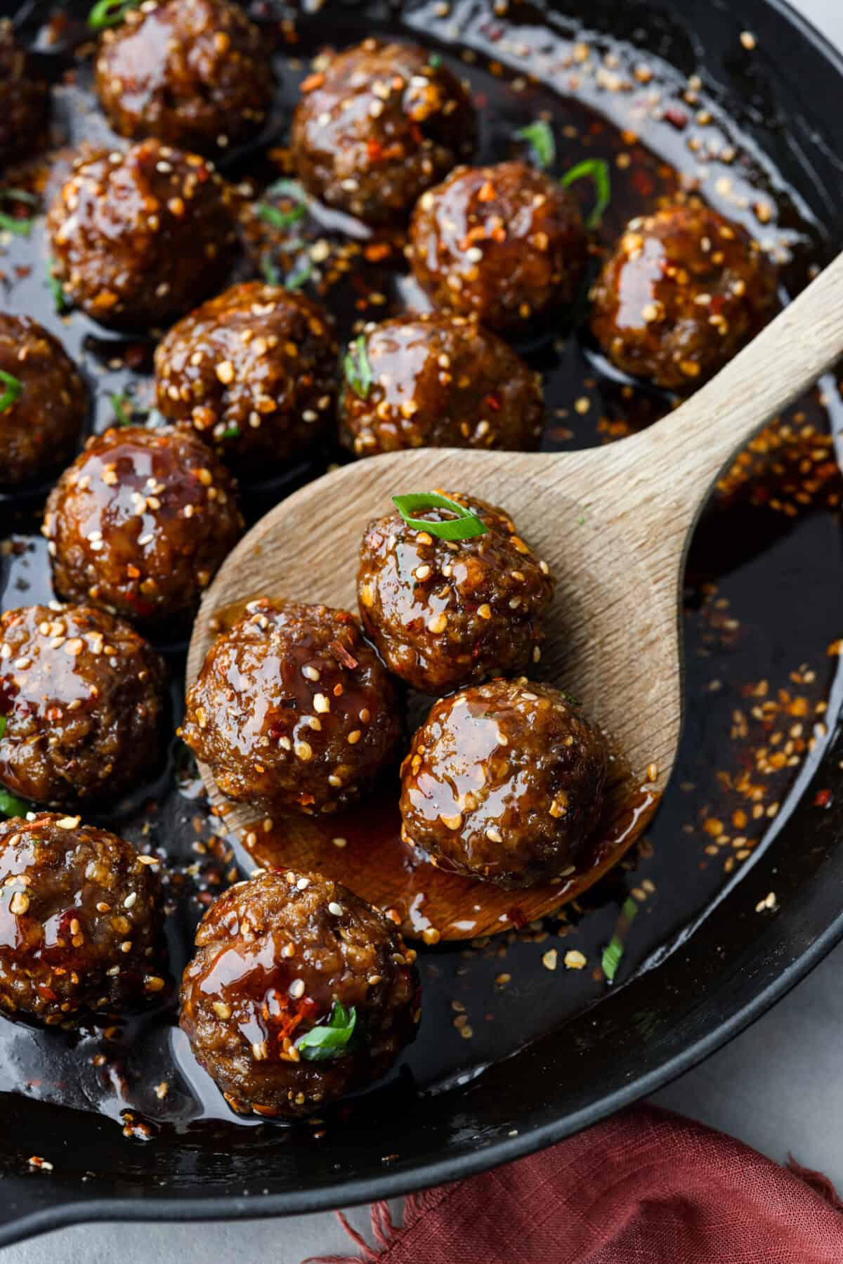Get ready to spice up your dinner with these firecracker meatballs! Juicy beef meatballs are baked to perfection and then tossed in a sweet and spicy buffalo-based sauce, creating a delicious meal that's perfect for any night of the week. Close up shot of large wooden spoon scooping meatballs out of a skillet.