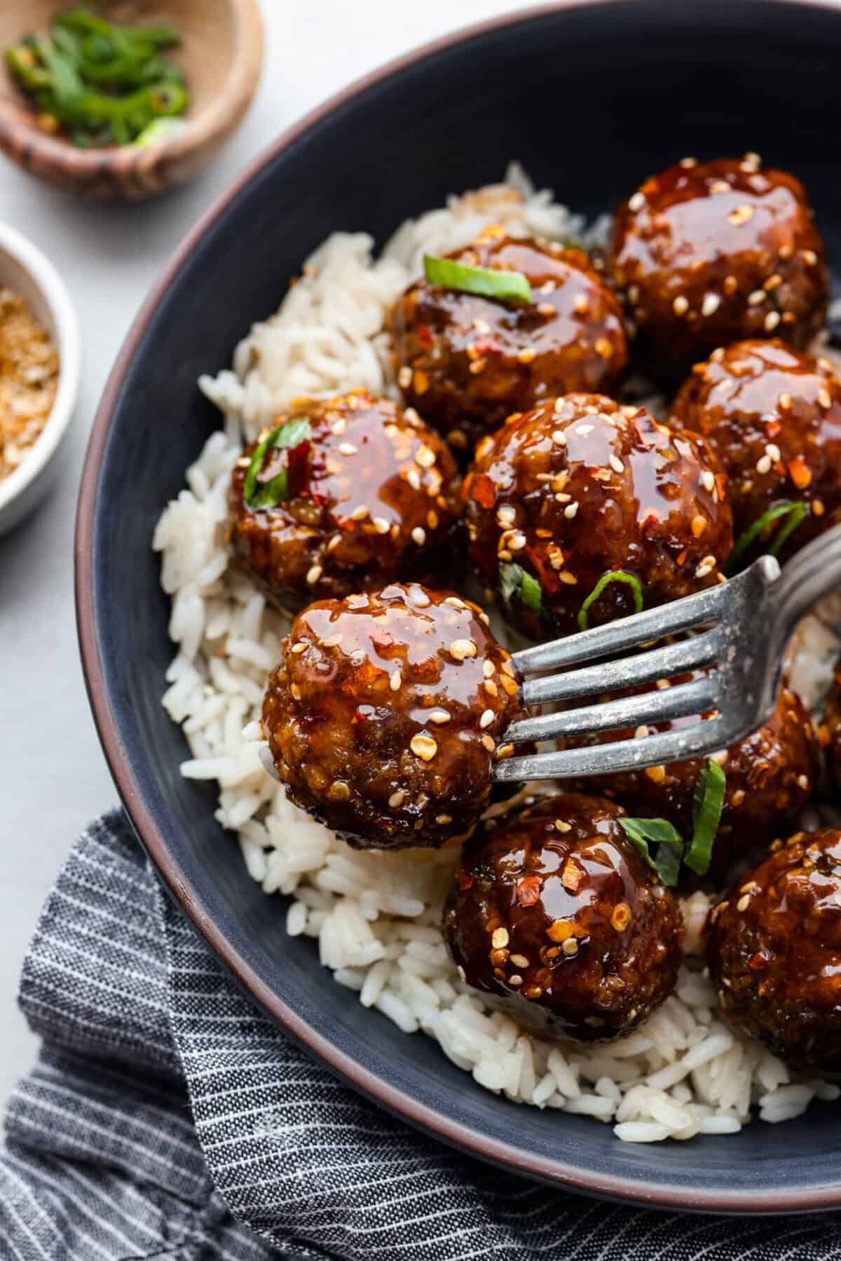 Get ready to spice up your dinner with these firecracker meatballs! Juicy beef meatballs are baked to perfection and then tossed in a sweet and spicy buffalo-based sauce, creating a delicious meal that's perfect for any night of the week. Close up shot of plated rice and a fork skewering a meatball.
