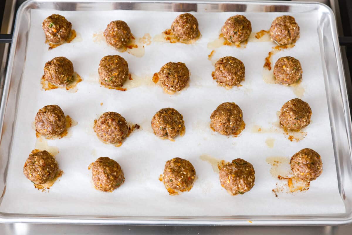 Get ready to spice up your dinner with these firecracker meatballs! Juicy beef meatballs are baked to perfection and then tossed in a sweet and spicy buffalo-based sauce, creating a delicious meal that's perfect for any night of the week. Overhead shot of parchment lined pan with rows of uncooked meatballs.