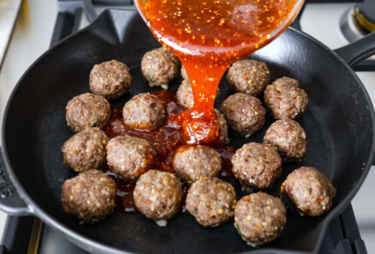 Get ready to spice up your dinner with these firecracker meatballs! Juicy beef meatballs are baked to perfection and then tossed in a sweet and spicy buffalo-based sauce, creating a delicious meal that's perfect for any night of the week. Angle shot of firecracker sauce being poured over cooked meatballs in a skillet.