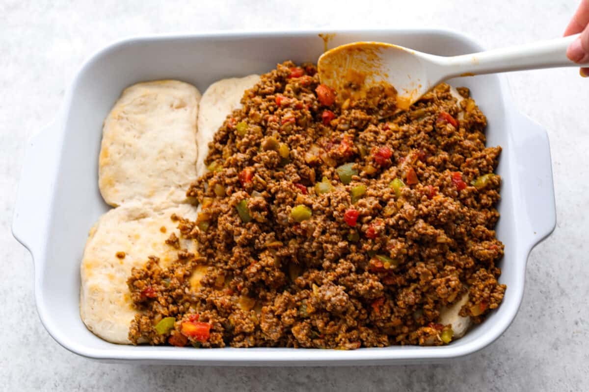 This delicious John Wayne Casserole is Mexican comfort food at its finest! Layers of seasoned ground beef, creamy cheddar cheese mixture, and more shredded cheese are baked on top of a biscuit crust for a crowd-pleasing family dinner! The meat mixture being added to the pressed biscuits.