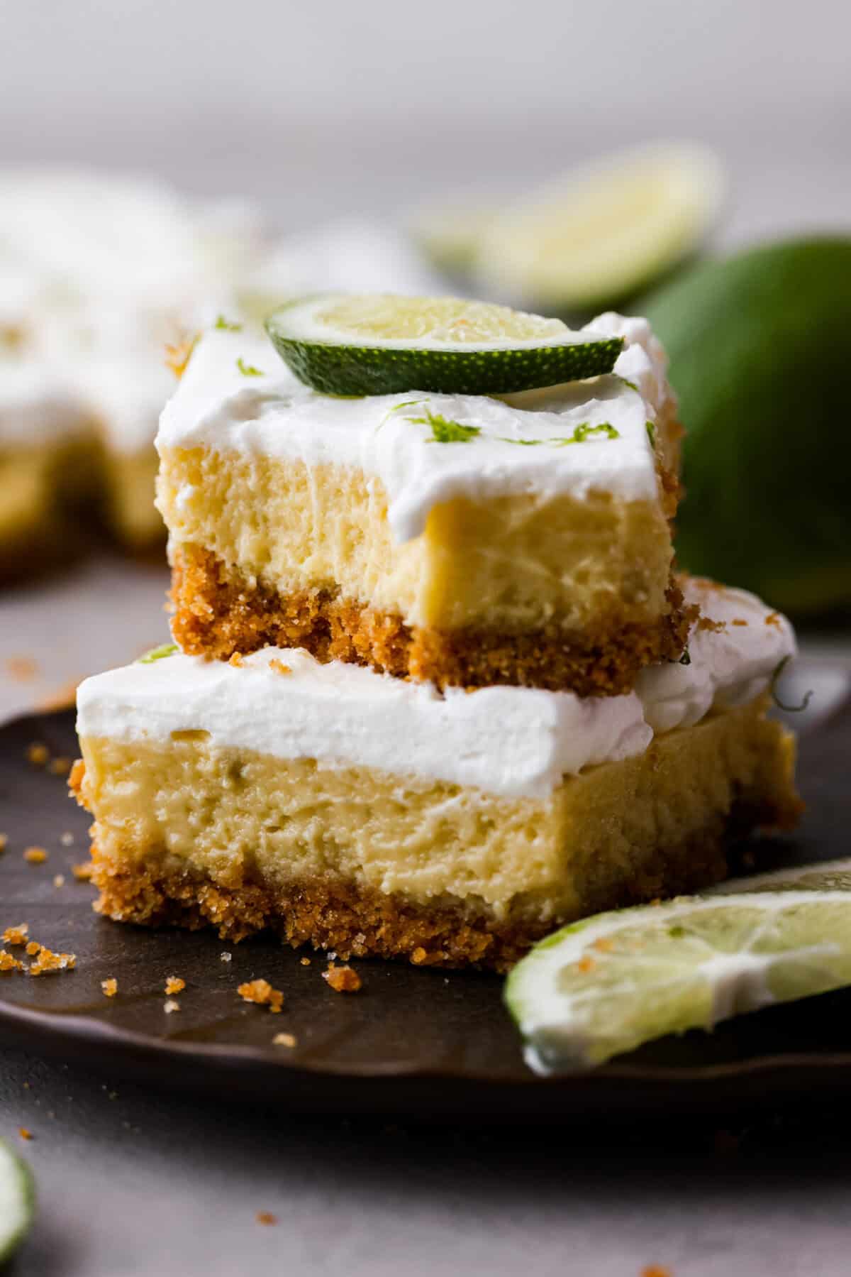 Key lime pie bars are classic key lime pie flavors baked into delightful little bars. They have a buttery graham cracker crust and a creamy lime filling. The sweet and zingy lime flavors make for an irresistible treat! Close view of key lime pie bars stacked on a brown plate.