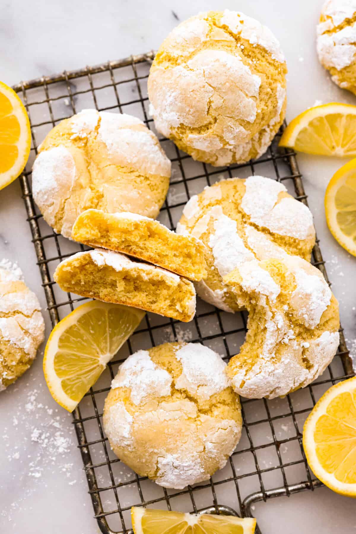 Lemon crinkle cookies are thick, soft pillows of lemon deliciousness! They have a refreshing flavor with an irresistible chewy texture. The perfect balance of sweet and tart make these a welcome treat for any occasion! The crinkle cookies on a wire rack. Some have a bite taken out of them or are broken in half.