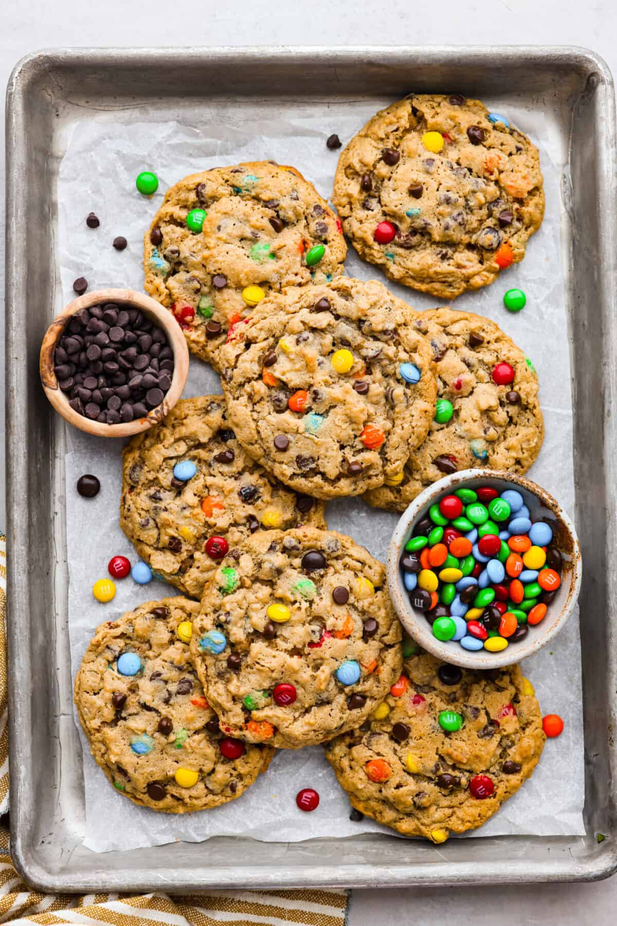 Monster cookies are slightly crisp on the outside, and soft and chewy in the center. They are loaded with peanut butter, oats, M&M candies, AND chocolate chips! Big monster cookies win every time! Top view of monster cookies on a sheet pan with bowls of M&M's and chocolate chips.