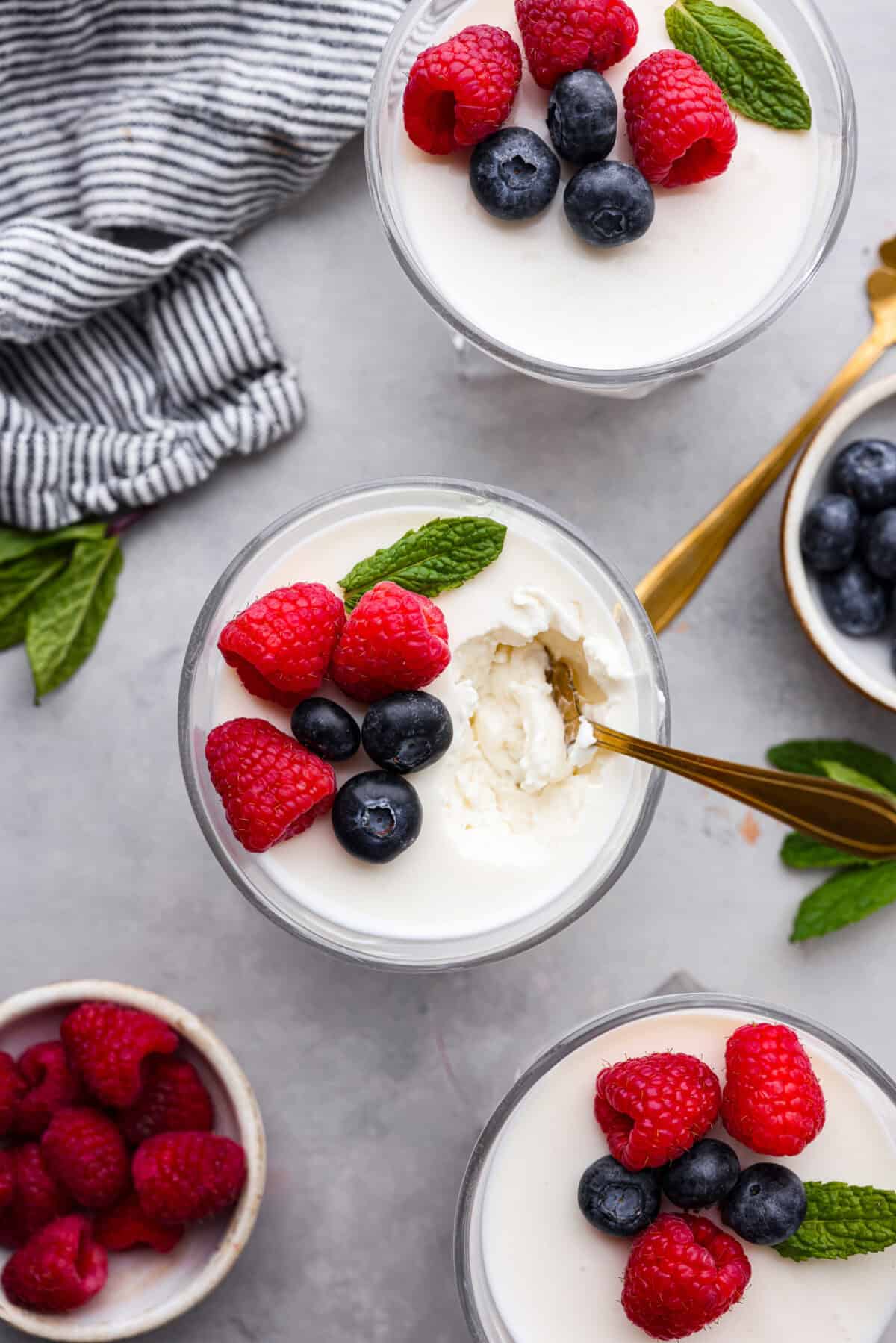 Treat yourself to this luxurious and refreshing Swedish Cream, a classic dessert that's easy to make. With a few simple ingredients and some chill time, you'll have a delightful treat topped with fresh berries and mint. Overhead shot of a cup of Swedish cream with berries and mint on top, but a gold spoon sticking out of the top. There are other bowls filled with raspberries and blueberries nearby.