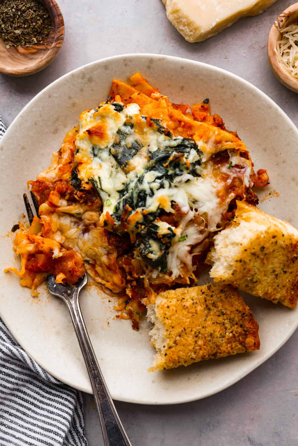 This super easy sheet pan lasagna is stacked with sautรฉedย ground beef and Italianย sausage that are cooked together, marinaraย sauce,ย creamy spinach ricotta, lasagna noodles and melty cheese for an amazingย Italian dinner. A small plate with a slice of lasagna and some garlic bread.