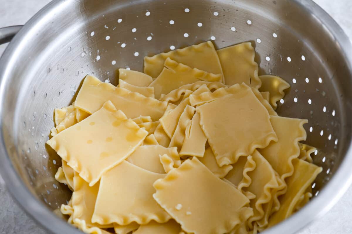 This super easy sheet pan lasagna is stacked with sautรฉedย ground beef and Italianย sausage that are cooked together, marinaraย sauce,ย creamy spinach ricotta, lasagna noodles and melty cheese for an amazingย Italian dinner. A strainer with lasagna noodles broken up.