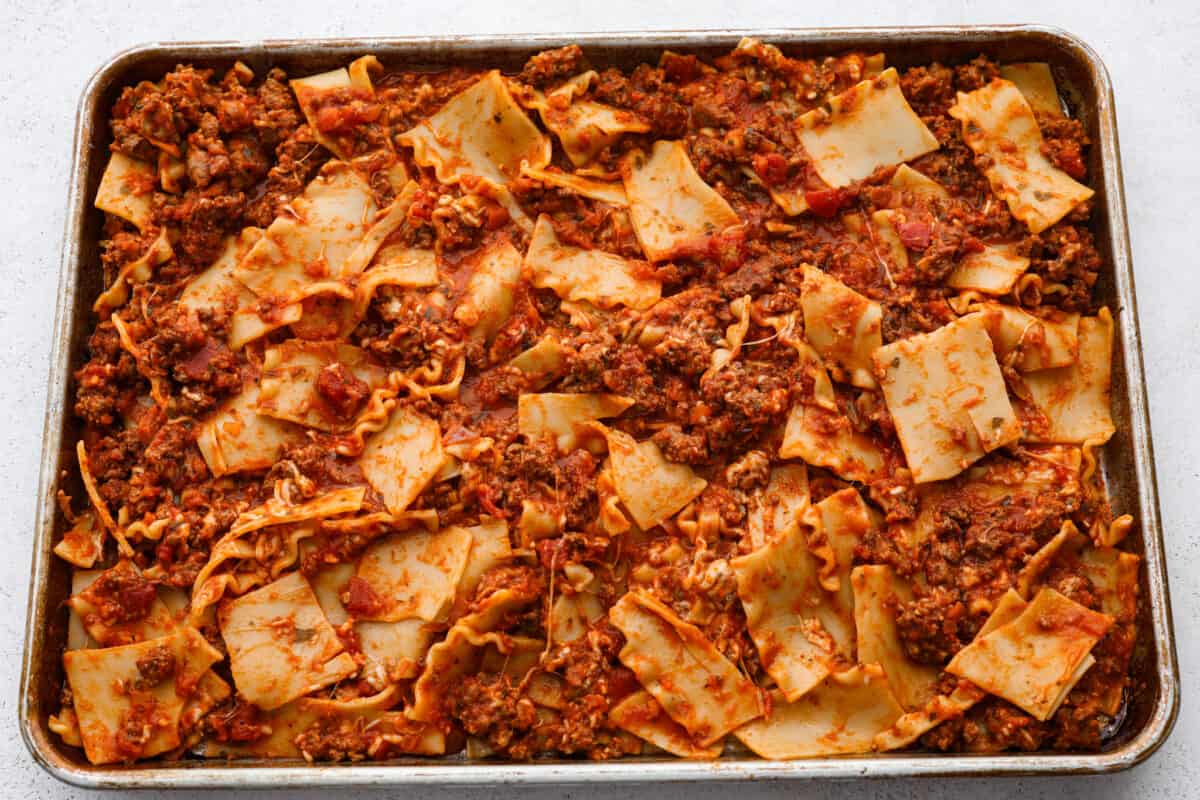This super easy sheet pan lasagna is stacked with sautรฉedย ground beef and Italianย sausage that are cooked together, marinaraย sauce,ย creamy spinach ricotta, lasagna noodles and melty cheese for an amazingย Italian dinner. Lasagna noodles and meat being spread on a sheet pan.
