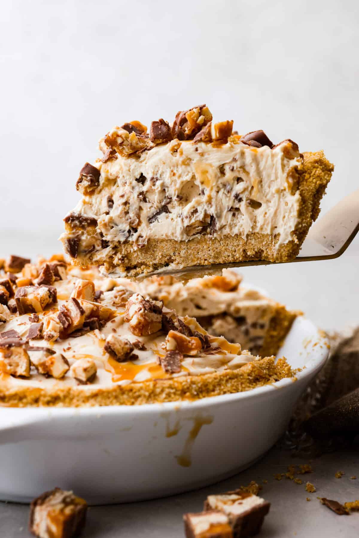 No Bake Snickers Bar Pie is one of the easiest and most delicious no bake pies that you make. You will love the Snickers pieces in every bite! A slice of no bake snickers bar pie being removed from the pie pan.