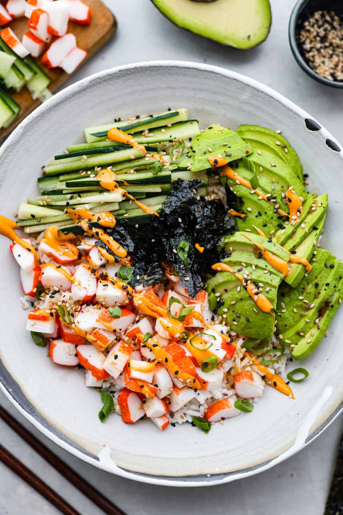 This sushi bowl is a deconstructed California sushi roll loaded with irresistible flavor and all of the best sushi ingredients. Itโs layered with sushi rice, crab, cucumber, avocado, a tasty sauce, and more. Itโs easy to make and ready to serve in just 20 minutes! Overhead shot of plated sushi bowl with sauce green onions and sesame seeds.