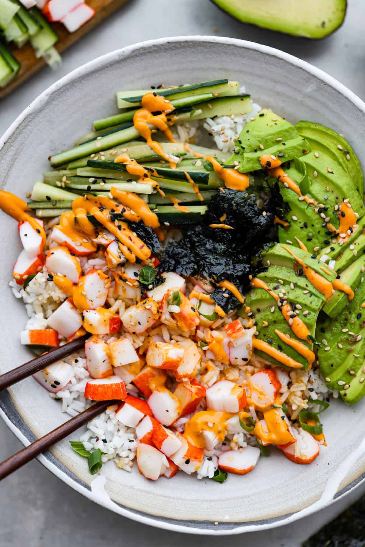 This sushi bowl is a deconstructed California sushi roll loaded with irresistible flavor and all of the best sushi ingredients. Itโs layered with sushi rice, crab, cucumber, avocado, a tasty sauce, and more. Itโs easy to make and ready to serve in just 20 minutes! Overhead shot of sushi bowl with chopsticks.