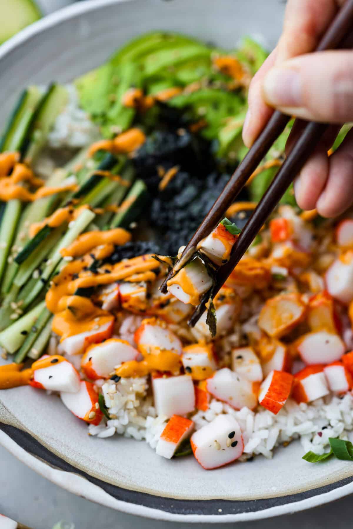 This sushi bowl is a deconstructed California sushi roll loaded with irresistible flavor and all of the best sushi ingredients. Itโs layered with sushi rice, crab, cucumber, avocado, a tasty sauce, and more. Itโs easy to make and ready to serve in just 20 minutes! Close up shot of someone taking a bite of the sushi bowl with chopsticks.