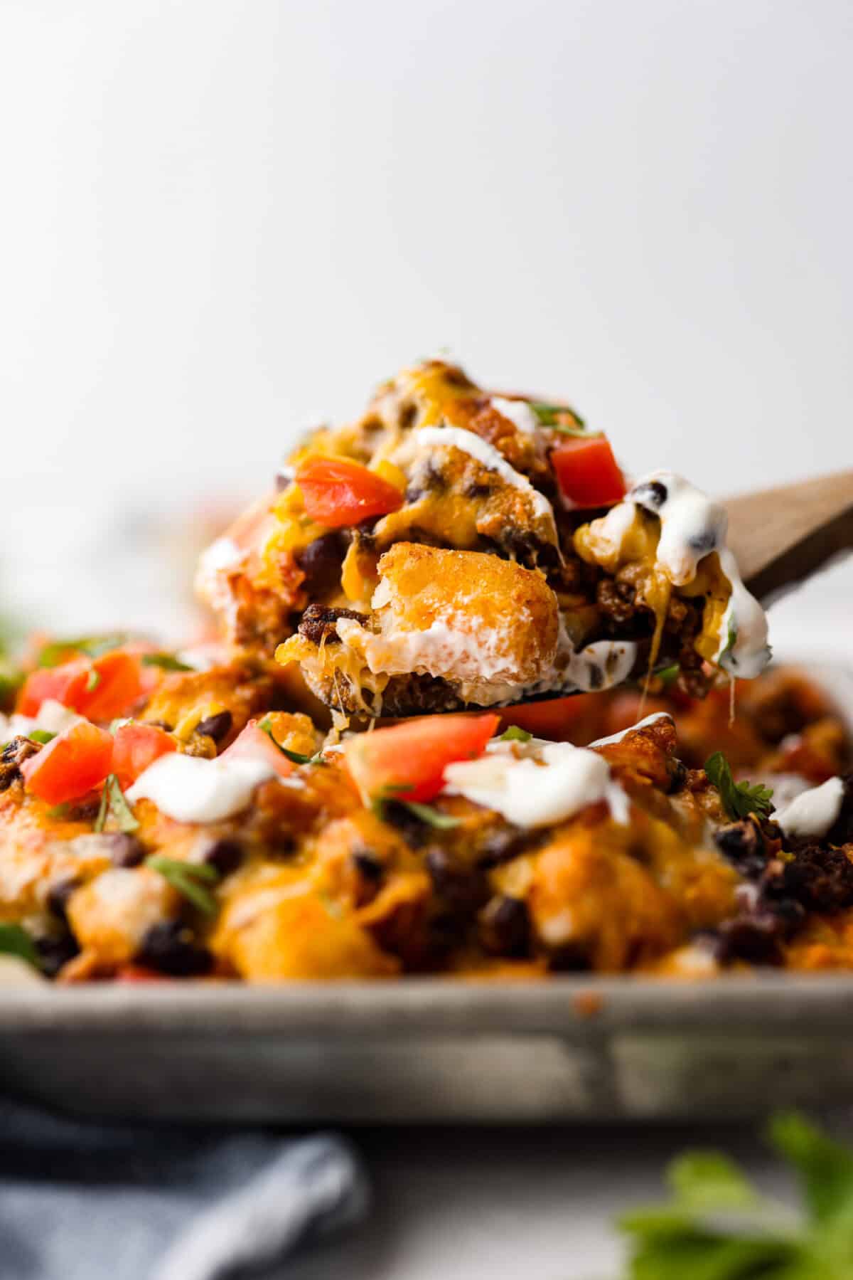 Tater Tot Nachos (Totchos) are made with crispy golden tater tots, topped with seasoned beef, scoops of refried beans, corn, black beans, salsa and melty cheese. Add your favorite toppings and dig in! Close up side shot of a spoon full of tater tot nachos being scooped out of the pan.