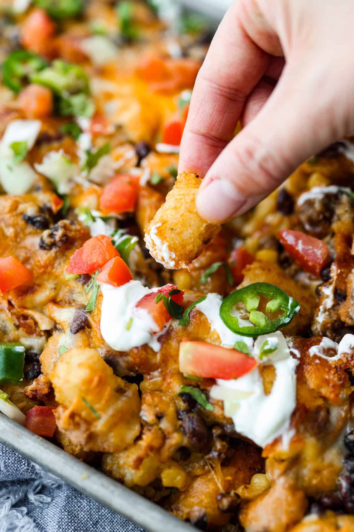 Tater Tot Nachos (Totchos) are made with crispy golden tater tots, topped with seasoned beef, scoops of refried beans, corn, black beans, salsa and melty cheese. Add your favorite toppings and dig in! Angle shot of someone dipping a tater tot into the sour cream on that is drizzled on top of the pan of totchos.