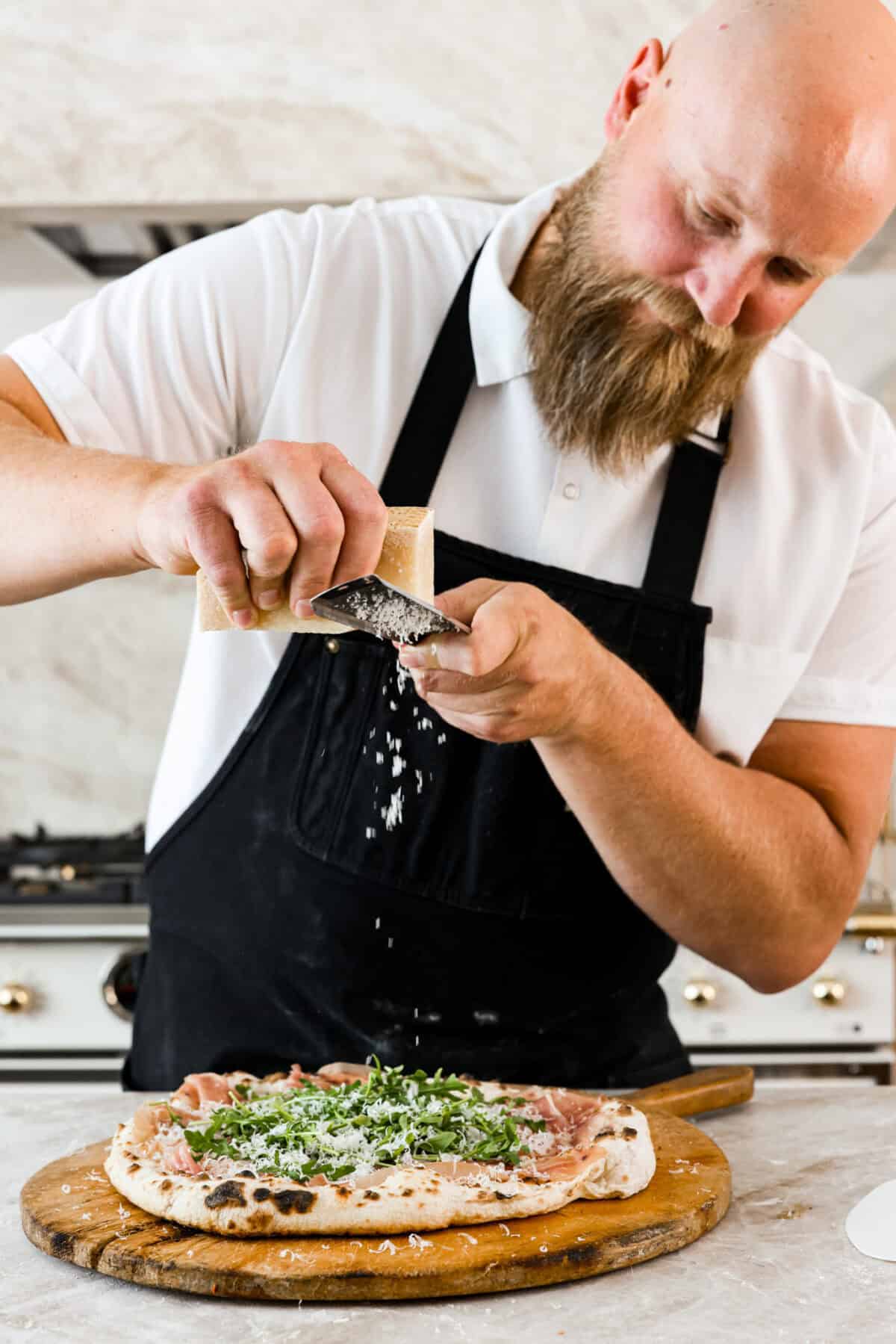 This is the perfect beginner's guide for making wood-fired pizza dough! Pizza expert, Chef Brandon Price, taught me how to make authentic homemade pizza, and now you get to learn too! Close view of the chef grating parmesan cheese on the wood-fired pizza.