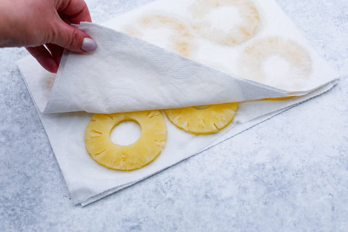 Perfect Pineapple Upside-Down Cake is a moist and fruity pineapple cake topped with caramelized pineapple rings and sweet maraschino cherries. It's no wonder this classic dessert has been a tried and true favorite for years! Third photo of the pineapple rings being pressed into a paper towel.