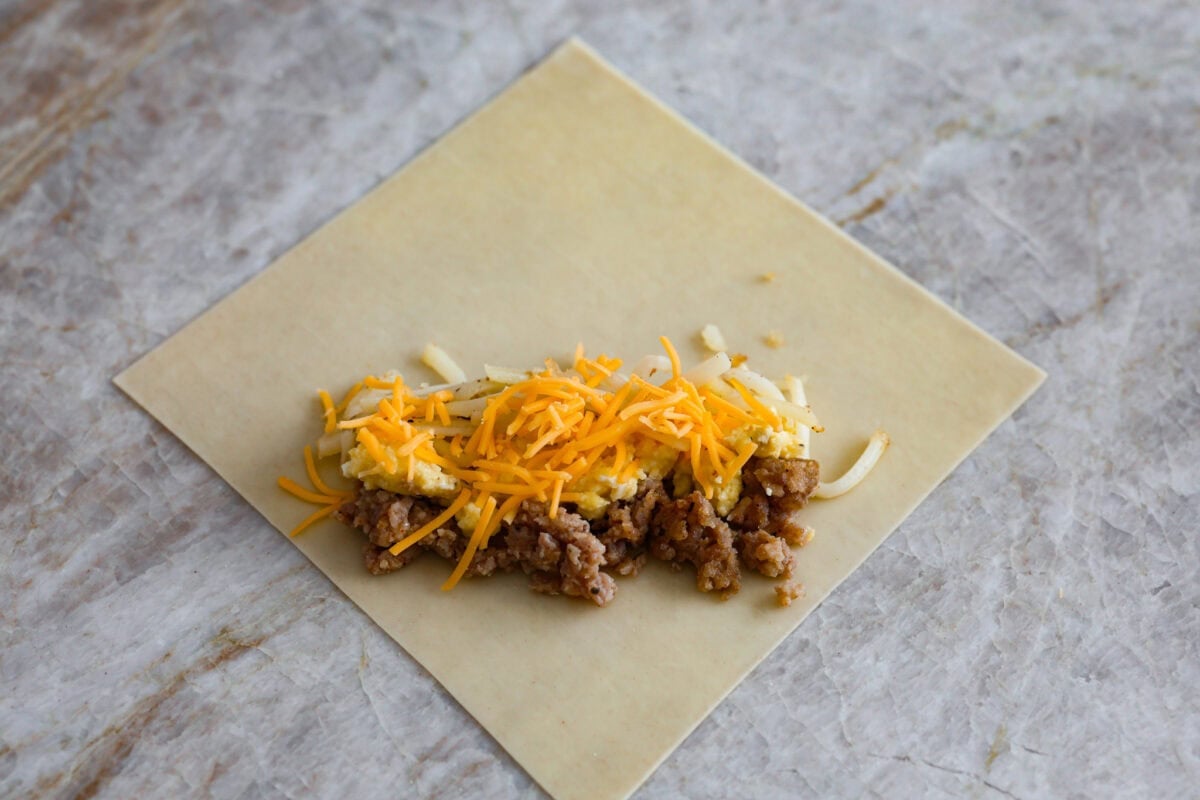 Breakfast Egg Rolls are a fun and delicious way to change up breakfast! The inside is filled with savory sausage, creamy eggs, hash browns, and plenty of cheese. Fry them up for a gooey, tender, savory center and a delicately crunchy exterior. This is a fun and unique breakfast that everyone will love! Overhead shot of egg roll paper open with ingredients for breakfast egg rolls on top.
