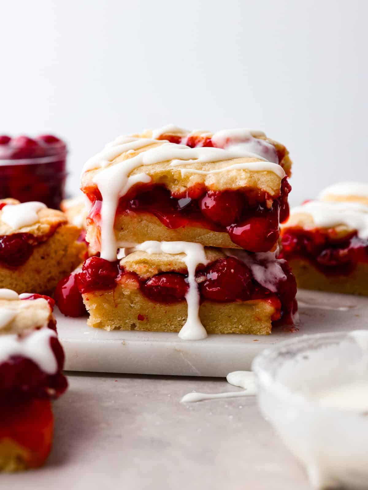 Enjoy cherry pie in its best form: cherry pie bars! You can whip up a whole pan in minutes using cherry pie filling. They’re beautiful, easy to make, and always a crowd-pleaser! A stack of cherry pie bars with the glaze drizzling off.