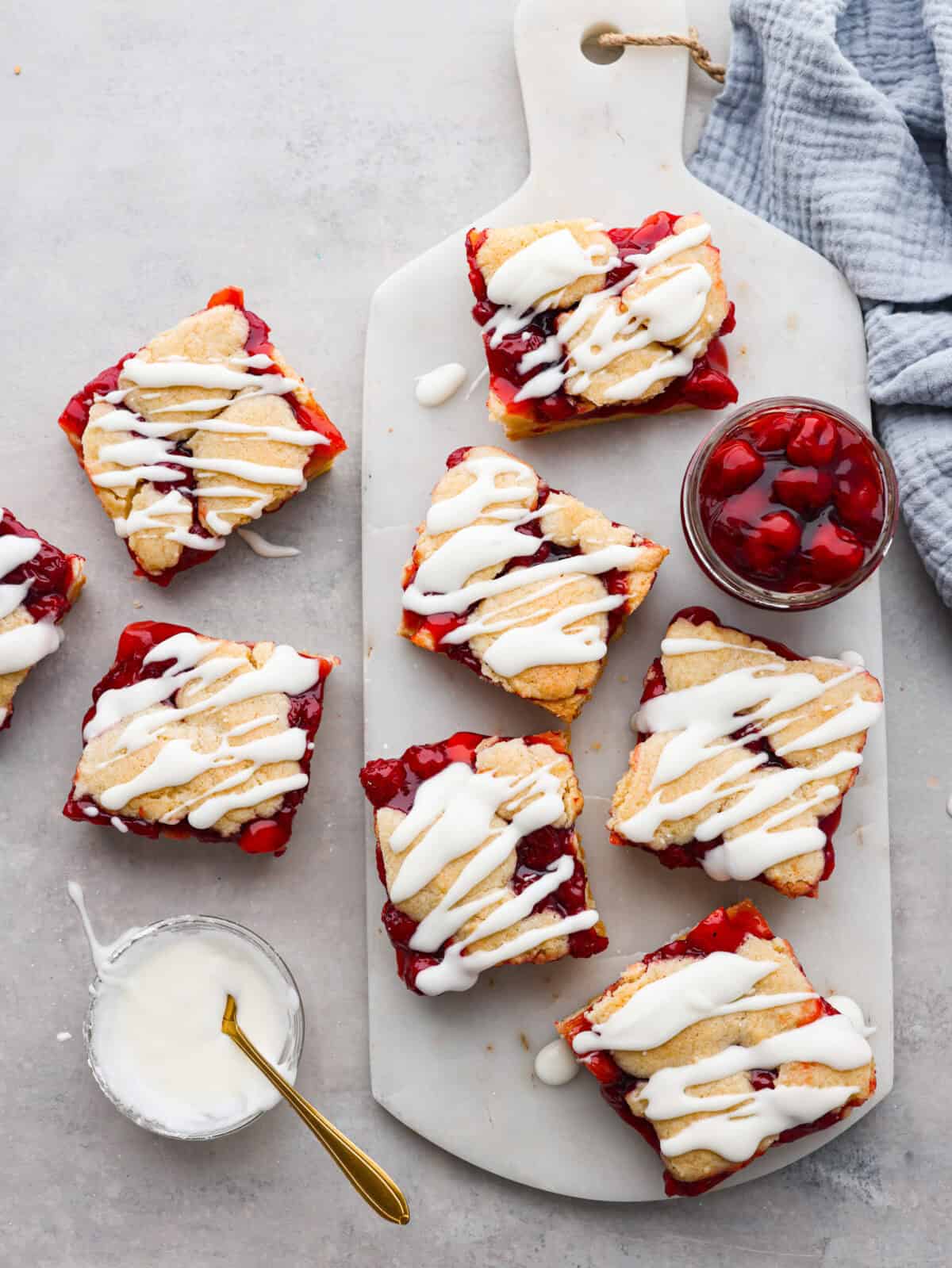 Enjoy cherry pie in its best form: cherry pie bars! You can whip up a whole pan in minutes using cherry pie filling. They’re beautiful, easy to make, and always a crowd-pleaser! A top view of baked bars on a cutting board. There is a bowl of glaze to the left with a gold spoon.