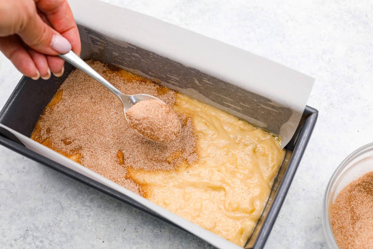 Cinnamon Swirl Banana Bread is a delightful twist on the classic. What sets this recipe apart is the luscious cinnamon sugar swirl running through the middle and on top, creating a beautiful marbled effect and an extra burst of flavor. It's irresistibly delicious! Overhead shot of banana bread batter in pan while someone sprinkles cinnamon and sugar mixture over the top.