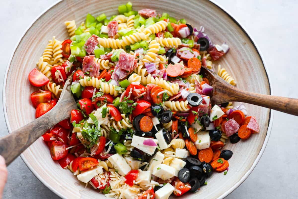 Indulge in the flavors of Italy with this irresistible Italian pasta salad! Each bite will be bursting with flavor from cherry tomatoes, creamy mozzarella, salami, olives, and fresh herbs topped with Italian dressing. It's like a taste of sunny Italy! Overhead shot of large bowl with two wooden spoons stirring ingredients of pasta salad together.