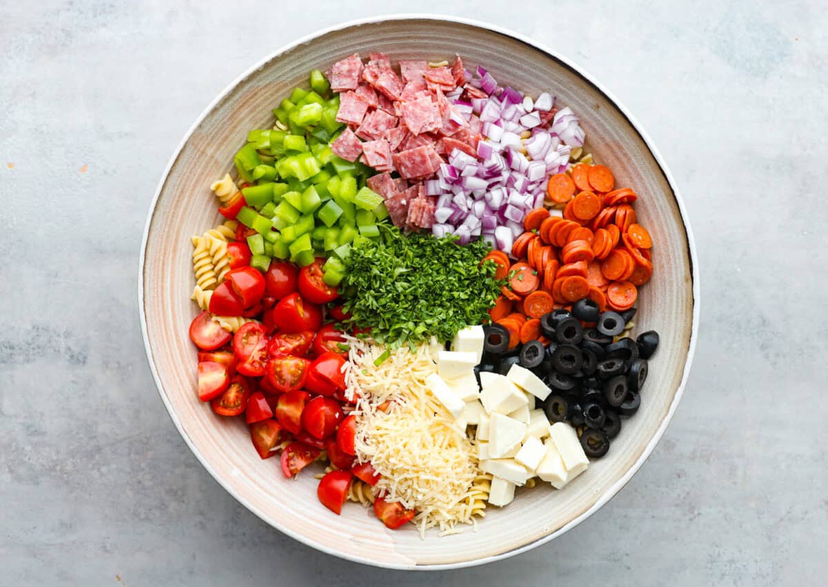 Indulge in the flavors of Italy with this irresistible Italian pasta salad! Each bite will be bursting with flavor from cherry tomatoes, creamy mozzarella, salami, olives, and fresh herbs topped with Italian dressing. It's like a taste of sunny Italy! Overhead shot of large bowl with ingredients separated into piles.