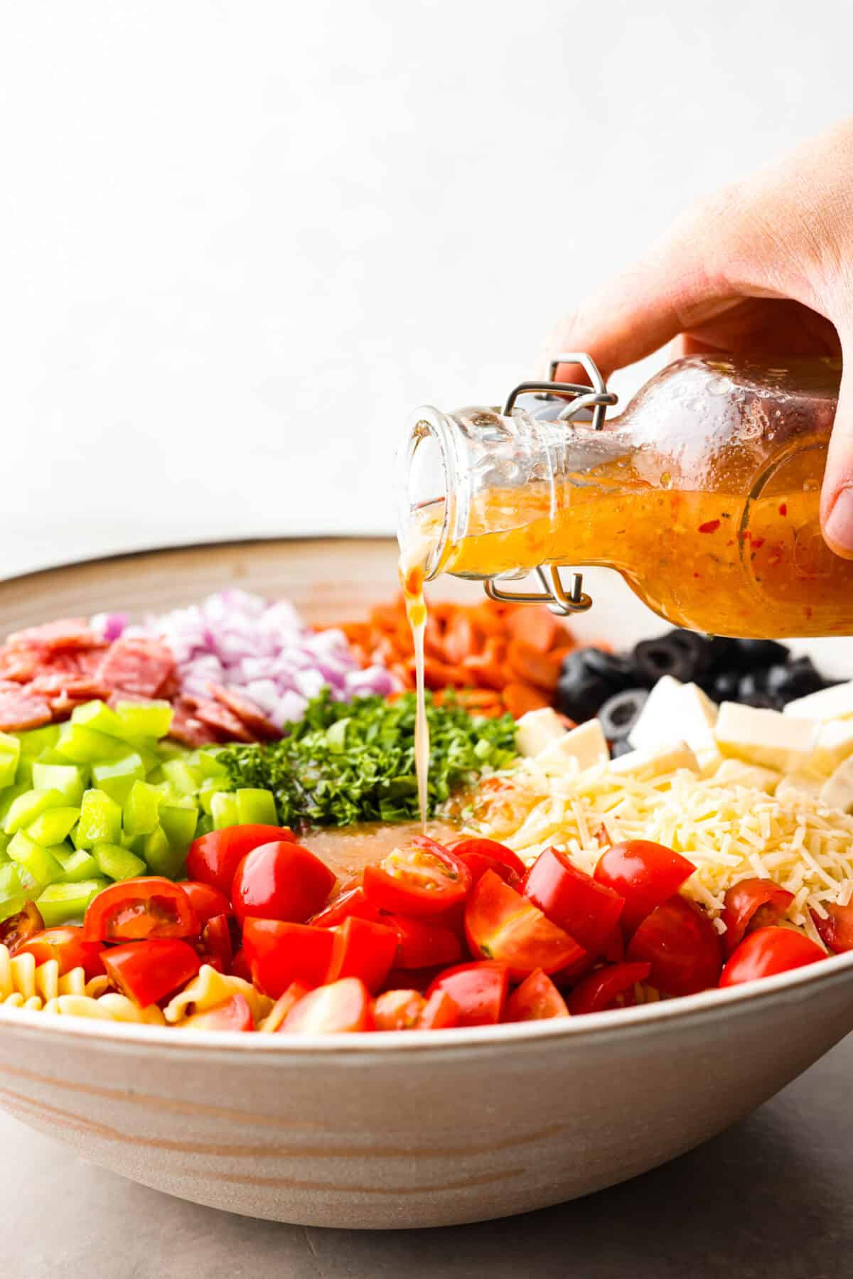 Indulge in the flavors of Italy with this irresistible Italian pasta salad! Each bite will be bursting with flavor from cherry tomatoes, creamy mozzarella, salami, olives, and fresh herbs topped with Italian dressing. It's like a taste of sunny Italy! Side view of large bowl with pasta salad ingredients in piles. Someone is pouring Italian dressing into the bowl.