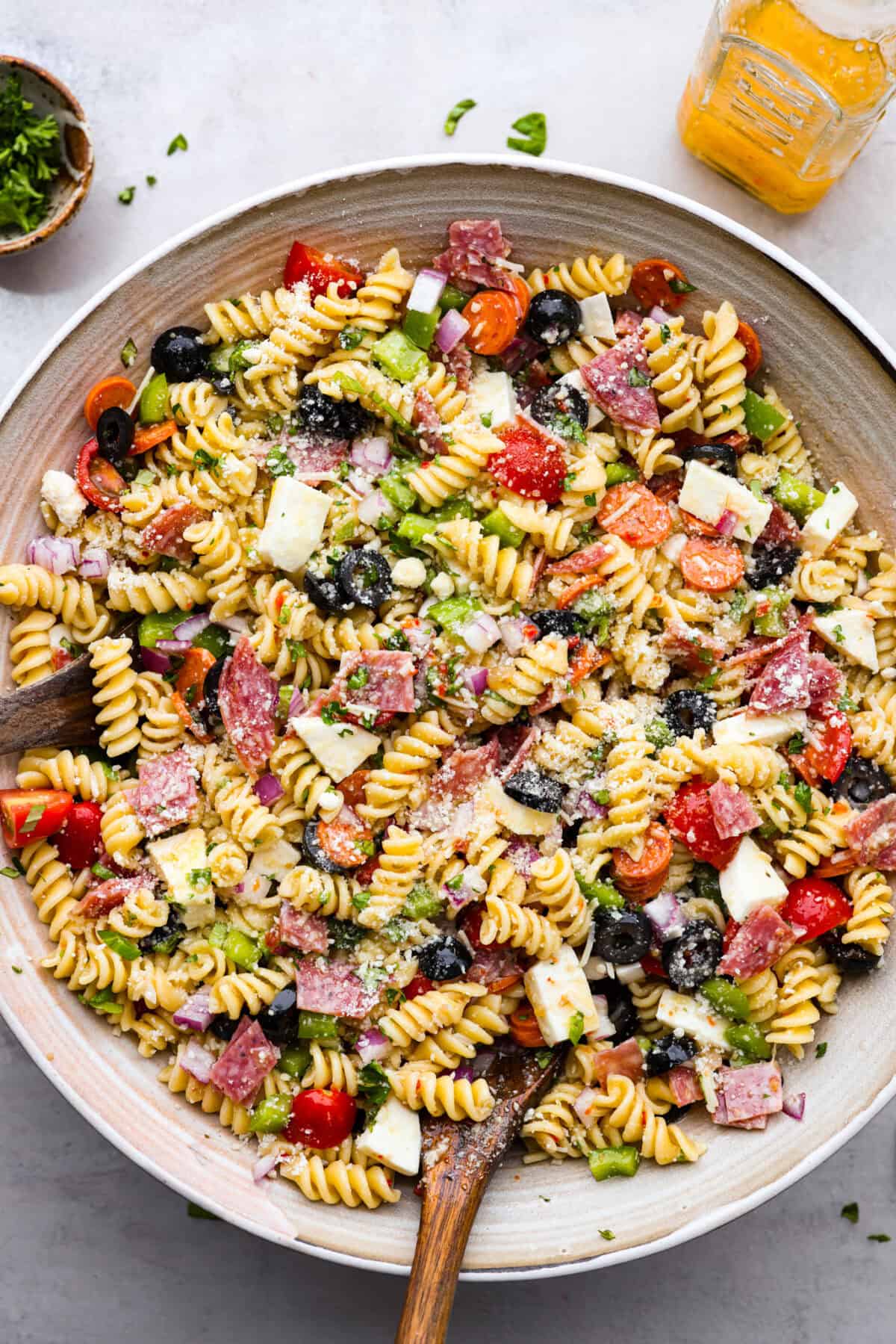 Indulge in the flavors of Italy with this irresistible Italian pasta salad! Each bite will be bursting with flavor from cherry tomatoes, creamy mozzarella, salami, olives, and fresh herbs topped with Italian dressing. It's like a taste of sunny Italy! Overhead shot of a large bowl of Italian pasta salad with wooden spoon sticking out