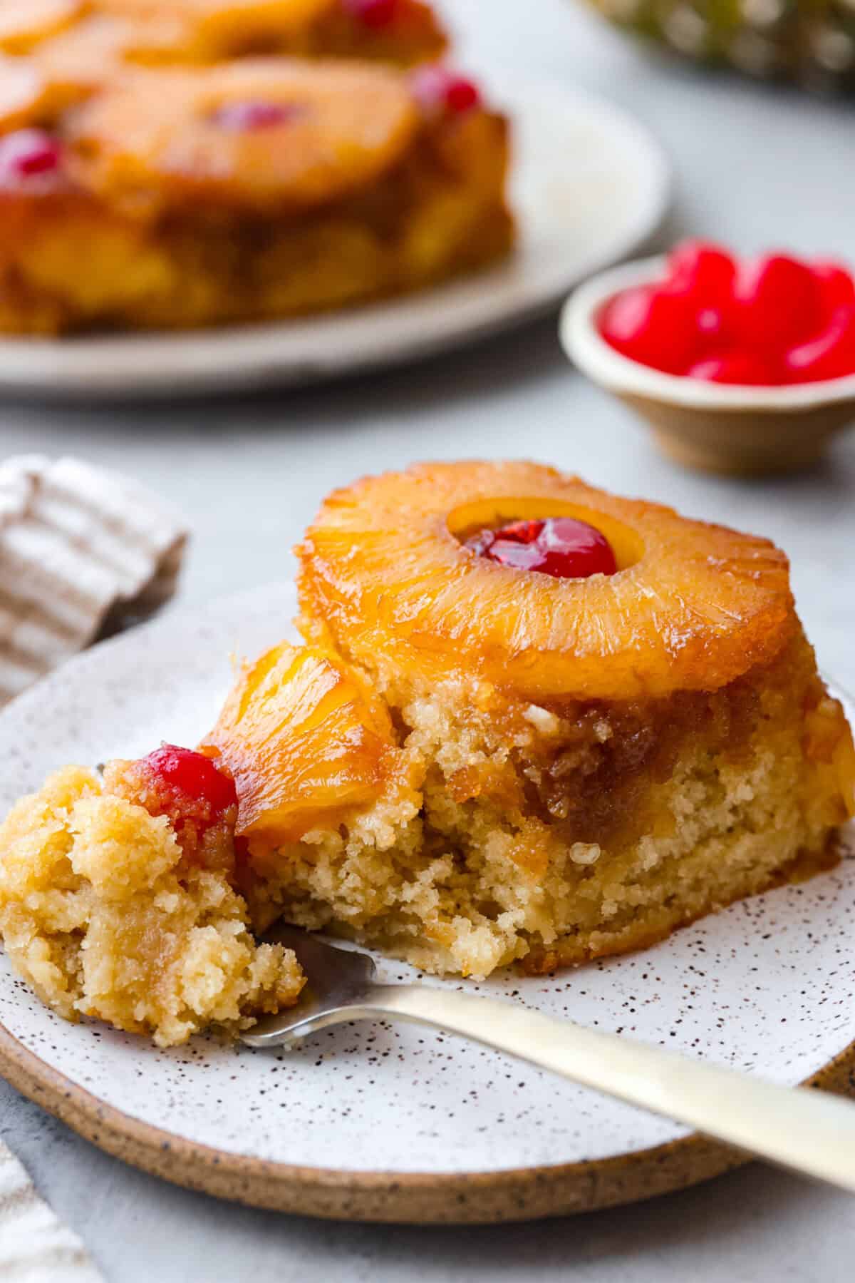 Perfect Pineapple Upside-Down Cake is a moist and fruity pineapple cake topped with caramelized pineapple rings and sweet maraschino cherries. It's no wonder this classic dessert has been a tried and true favorite for years! Close view of a slice of pineapple upside down cake on a plate with a fork.