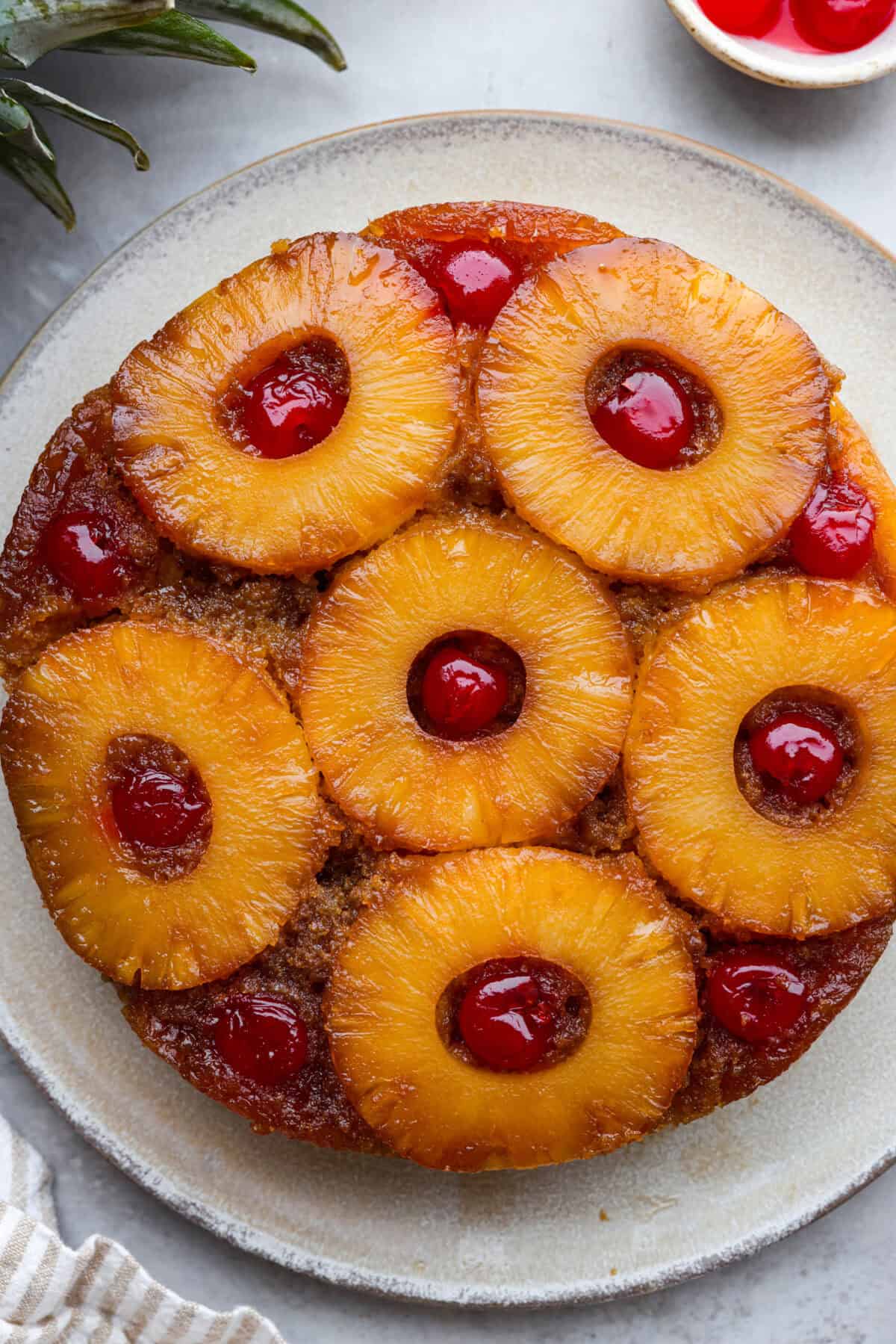 Perfect Pineapple Upside-Down Cake is a moist and fruity pineapple cake topped with caramelized pineapple rings and sweet maraschino cherries. It's no wonder this classic dessert has been a tried and true favorite for years! Top view of pineapple upside down cake on a plate.