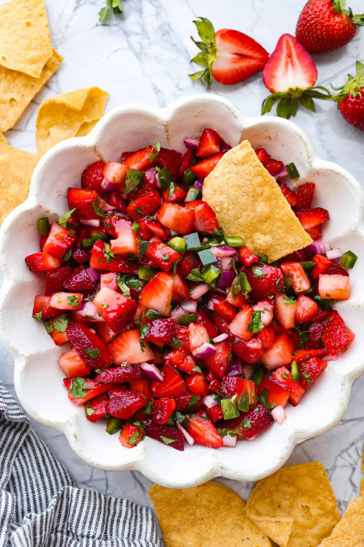 Add a burst of fresh, fruity flavor to your table with this Strawberry Salsa. This quick and easy recipe combines sweet strawberries with a hint of spice and is perfect for a light and refreshing snack in just 10 minutes! Top view of strawberry salsa in a scalloped bowl with a tortilla chip in it.