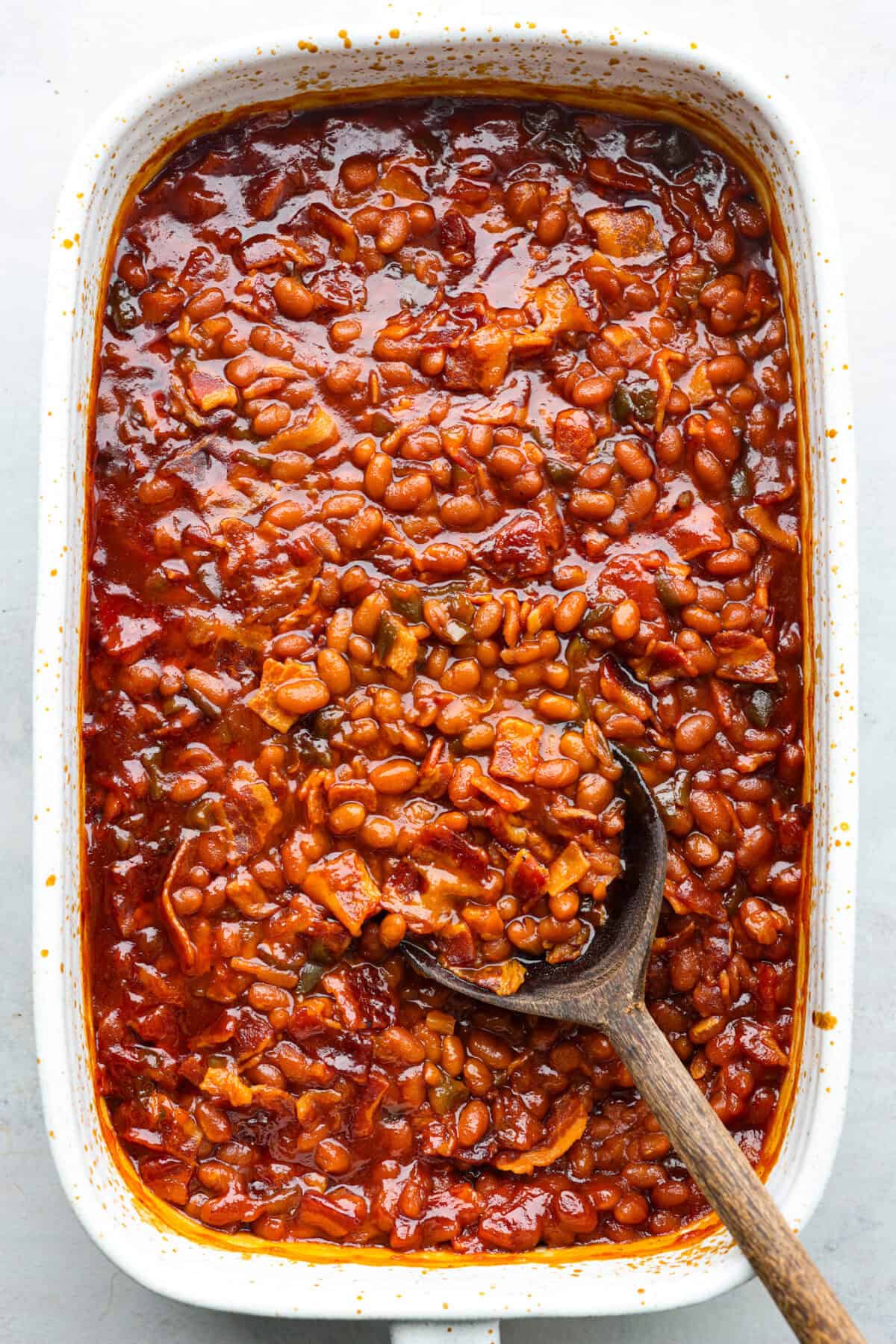 These World's Best Baked Beans will be the last recipe you will ever make! This easy recipe is the perfect side to a potluck and a tried and true family recipe that your family will go crazy over! Top view of the world's best baked beans in a baking dish with a wood spoon.