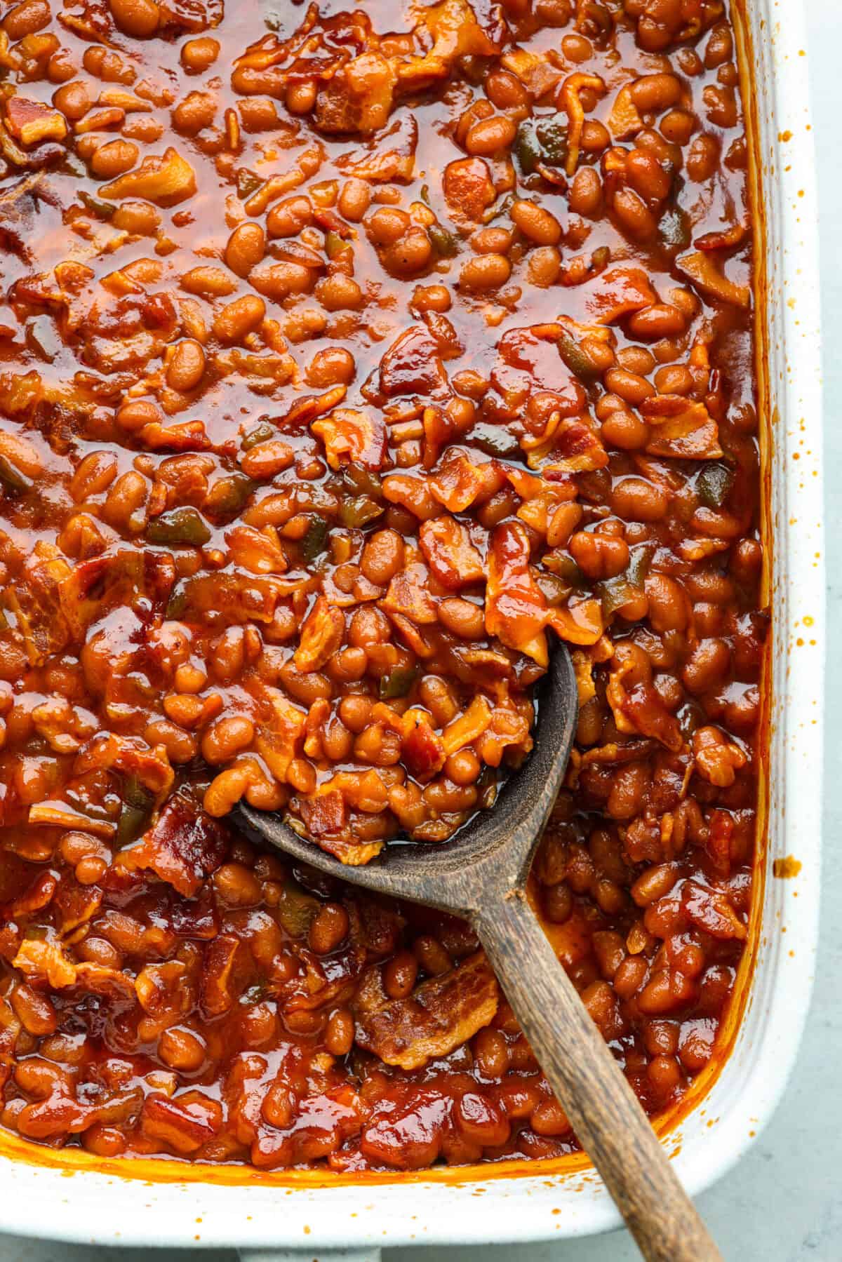 These World's Best Baked Beans will be the last recipe you will ever make! This easy recipe is the perfect side to a potluck and a tried and true family recipe that your family will go crazy over! Close view of the baked beans in a baking dish with a wooden spoon.
