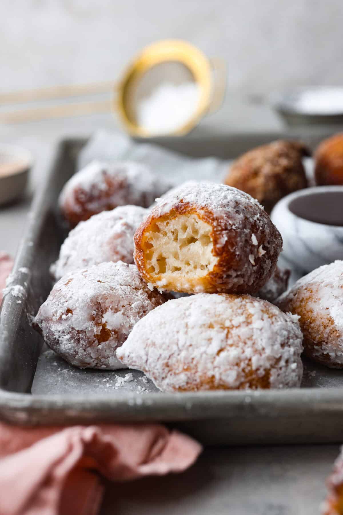 Whip up a batch of these delightful zeppole, perfect for a sweet treat anytime! With a light, fluffy texture and a generous coating of powdered or cinnamon sugar, these Italian donuts are sure to please. Side shot of zeppole coated in powdered sugar, one with a bite taken out of it.