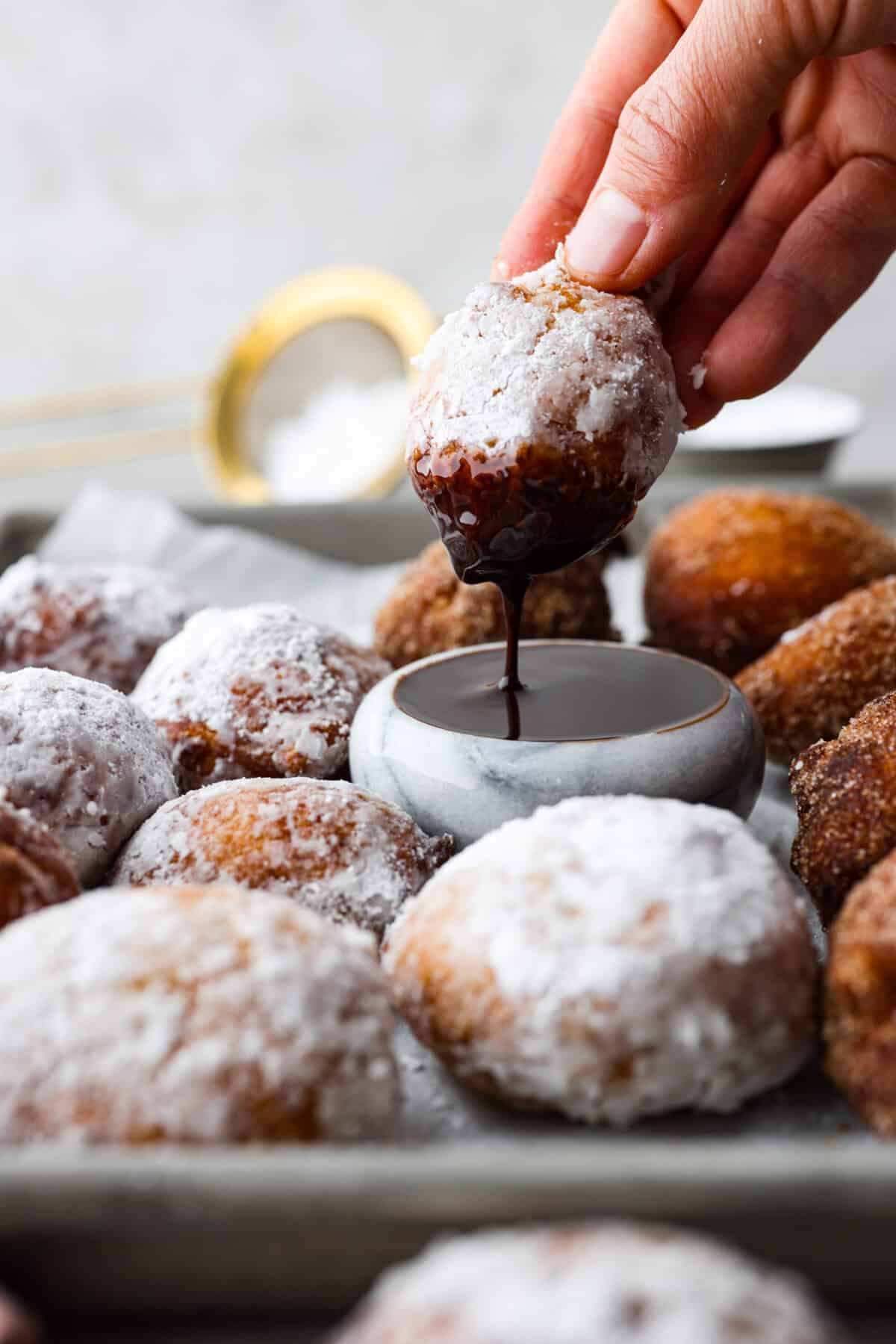 Whip up a batch of these delightful zeppole, perfect for a sweet treat anytime! With a light, fluffy texture and a generous coating of powdered or cinnamon sugar, these Italian donuts are sure to please. Side shot of someone dipping a zeppole in to chocolate sauce.