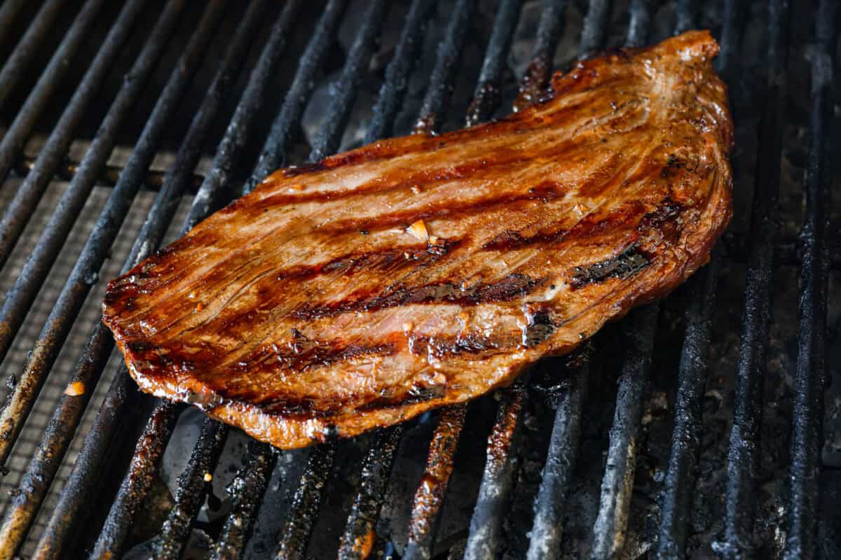 This grilled flank steak with Asian-inspired marinade features a savory blend of soy sauce, ginger, and garlic. The steak is seared to perfection on a hot grill, locking in all the delicious flavors. Best of all, itโs ready in less than 10 minutes! Angle shot of cooked steak on grill.