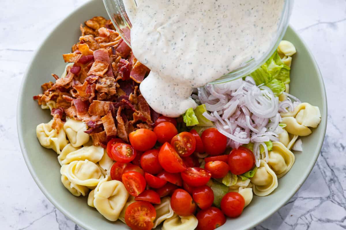 BLT Tortellini Pasta Salad is your favorite sandwich turned into a delicious, refreshing salad you can share with others. This salad is so easy to make and perfect for serving at any gathering. With loads of bacon, crisp Romaine lettuce, juicy tomatoes, and cheese-filled tortellini, it's sure to be a hit! Overhead shot of dressing being poured over salad.