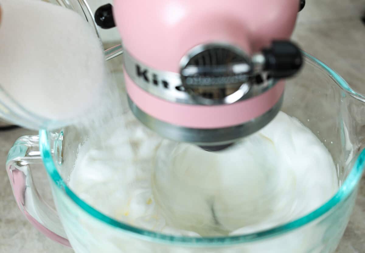 Impress everyone with this gorgeous Baked Alaska dessert! It's surprisingly easy to make with just 5 ingredients. Layers of cake, ice cream and meringue will have your friends and family begging for a slice! Angle shot of sugar being poured into stand mixer bowl full of whipped egg whites.