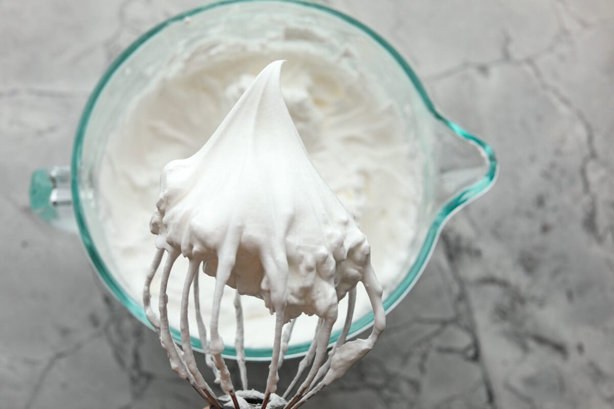 Impress everyone with this gorgeous Baked Alaska dessert! It's surprisingly easy to make with just 5 ingredients. Layers of cake, ice cream and meringue will have your friends and family begging for a slice! Overhead shot with bowl of whipped egg whites in the background with a stand mixer whisk attachment in the foreground with peaked egg whites.