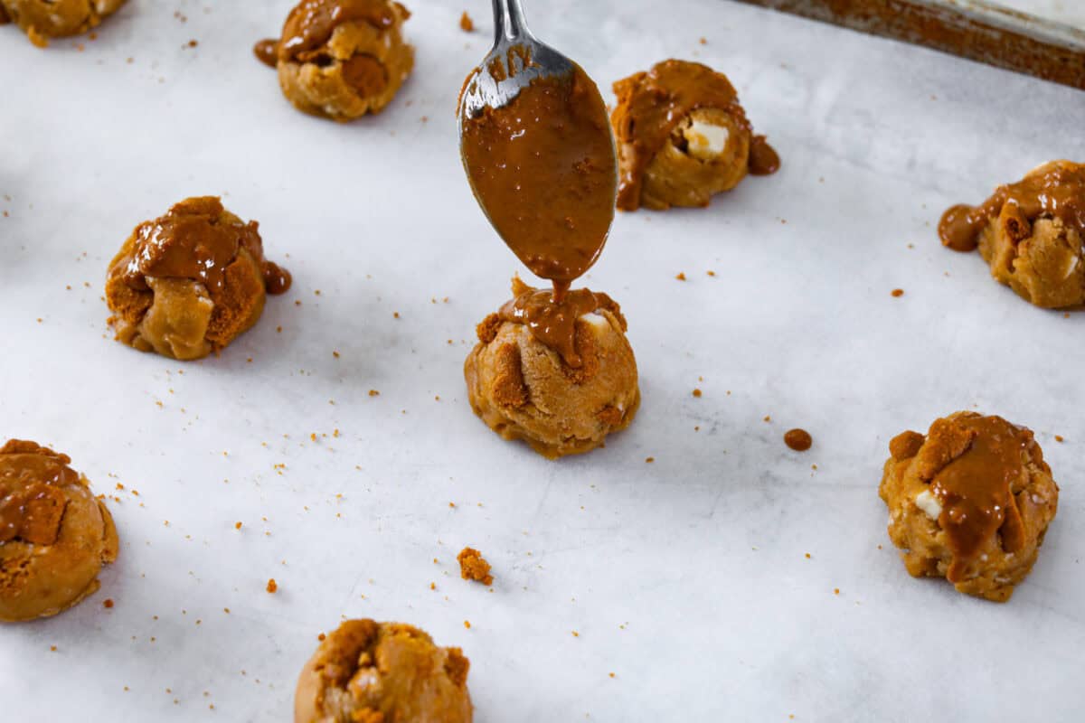 Satisfy your sweet tooth with loaded Biscoff Cookies. They are soft and chewy and infused with the flavor of Biscoff cookies and cookie butter. These are easy to whip up and will be a hit for any occasion! Angle shot of a spoon drizzling melted cookie butter over cookie dough balls on cookie sheet.