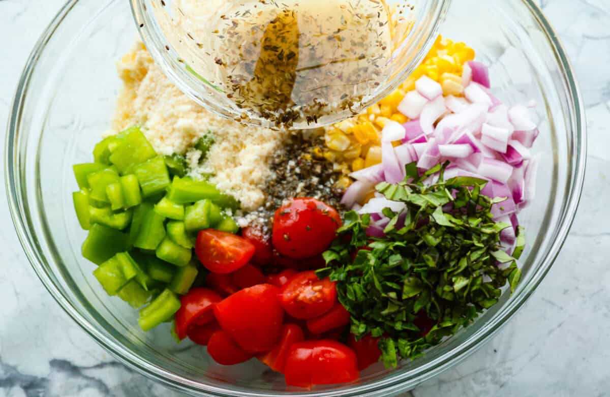 This Italian Corn Salad is bursting with fresh flavors from sweet corn, juicy grape tomatoes, crisp green bell pepper, and sharp parmesan cheese. Tossed in a zesty homemade Italian dressing, it’s perfect for summer or any time of the year. Overhead shot of dressing being poured into bowl of separated salad ingredients.