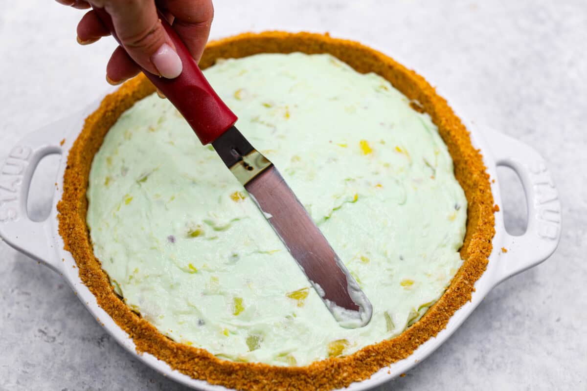 Bring this easy and irresistible no-bake pistachio cream pie to your next gathering! You'll love the combination of the pistachio and pineapple flavors that fill the inside of a sweet, buttery, graham cracker crust. Third photo of a small spatula smoothing the top of the no-bake pistachio cream pie.