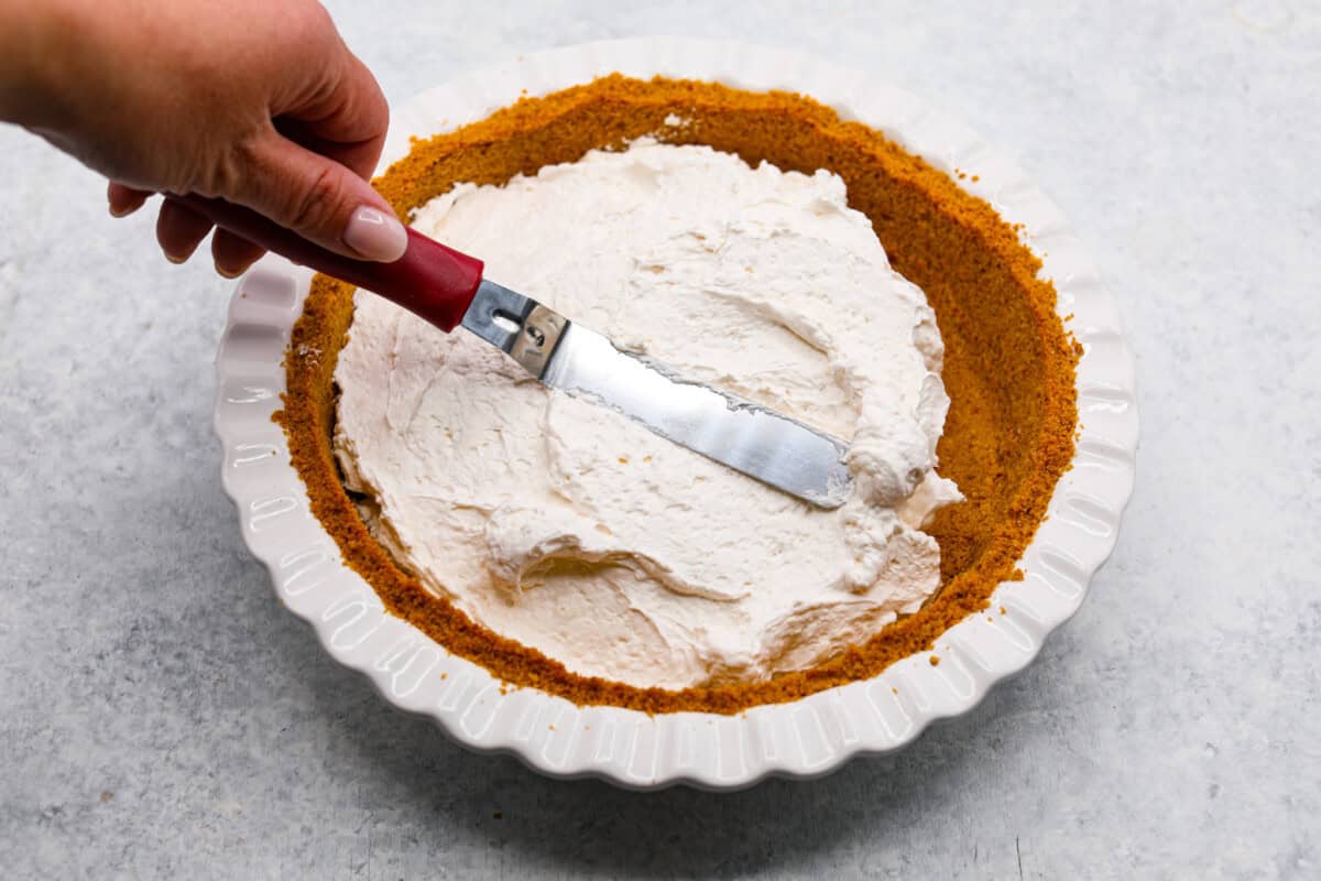 This strawberry cream cheese pie has a smooth, creamy filling is topped with sweet strawberries and is held together by a crumbly, buttery crust. Simply divine! Overhead shot of someone smoothing pie filling with metal spatula into graham cracker crust.