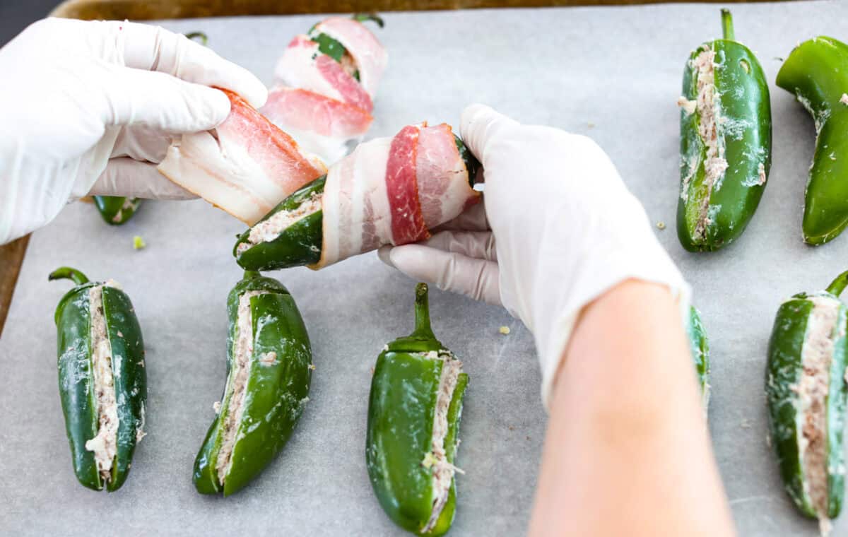 Texas Twinkies are a major upgrade to your classic jalapeรฑo poppers! They're jalapeรฑos filled with cream cheese, brisket, and shredded cheese, then wrapped in bacon and cooked to perfection. These are the hit of any party! Overhead shot of someone wrapping stuffed peppers with bacon.