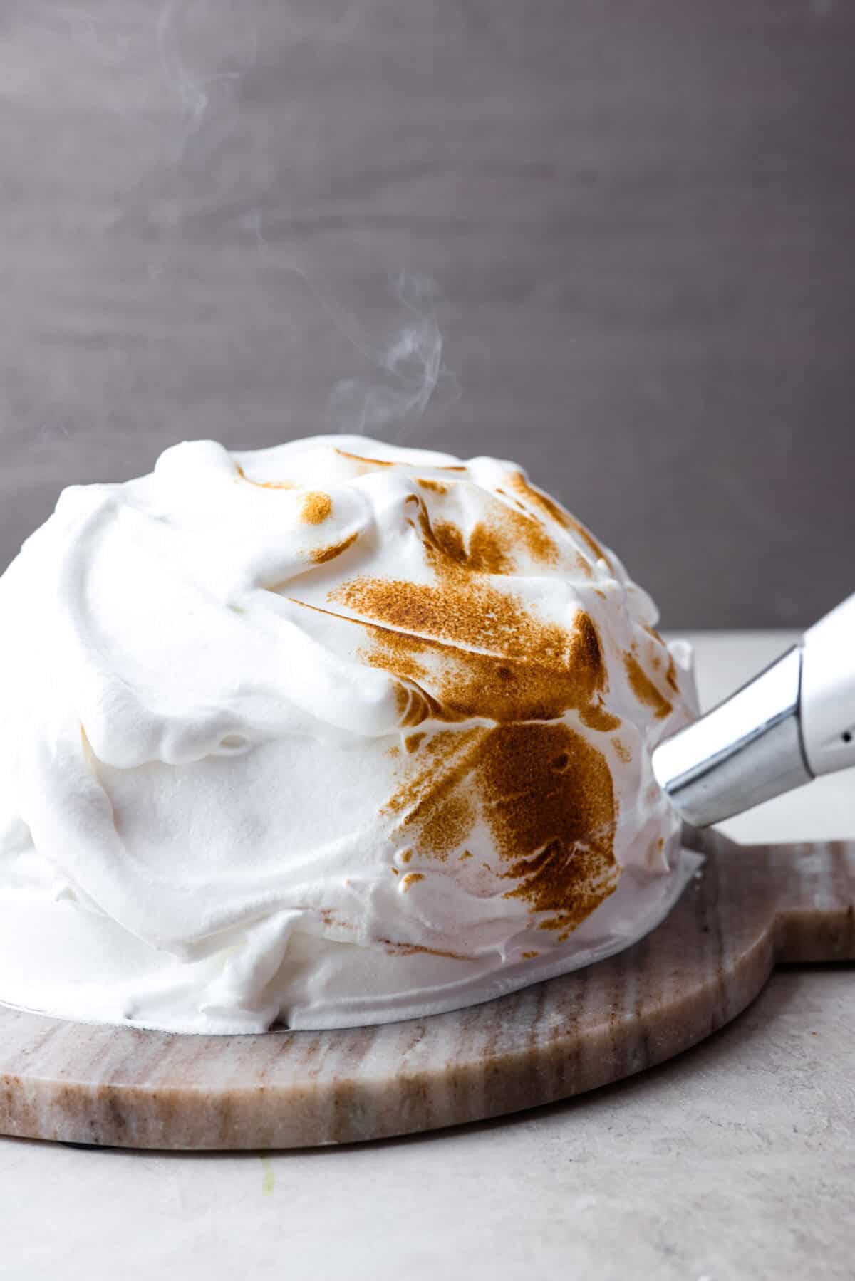 Impress everyone with this gorgeous Baked Alaska dessert! It's surprisingly easy to make with just 5 ingredients. Layers of cake, ice cream and meringue will have your friends and family begging for a slice! Side shot of baked Alaska covered in meringue being browned with a hand torch.