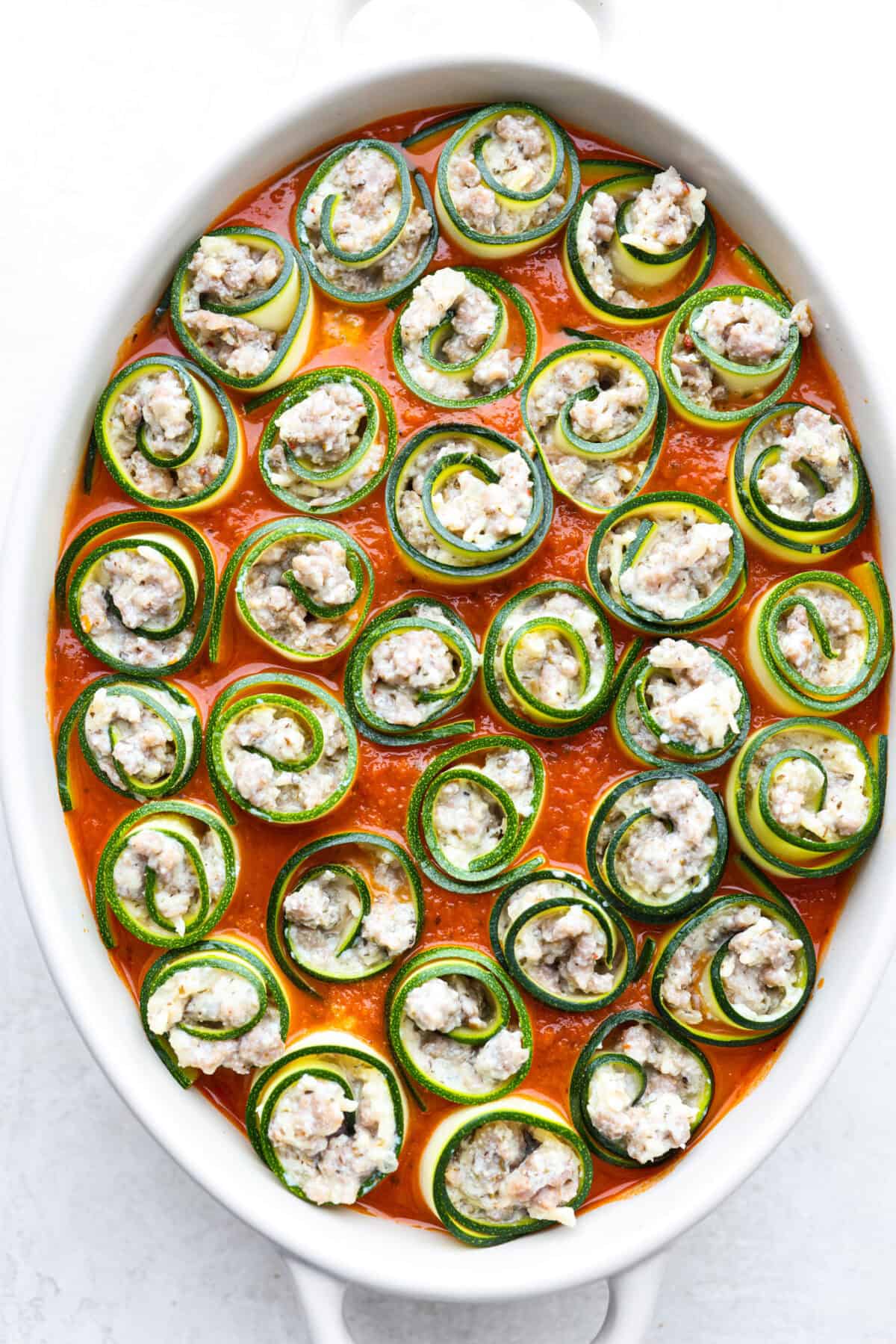 Zucchini Roll-Ups are a delicious, unique meal that the whole family will enjoy. Thin slices of zucchini are rolled around a tasty sausage ricotta mixture and layered over marinara before being topped with cheese and baked to bubbly perfection! Overhead shot of zucchini rolls in marinara sauce in pan.
