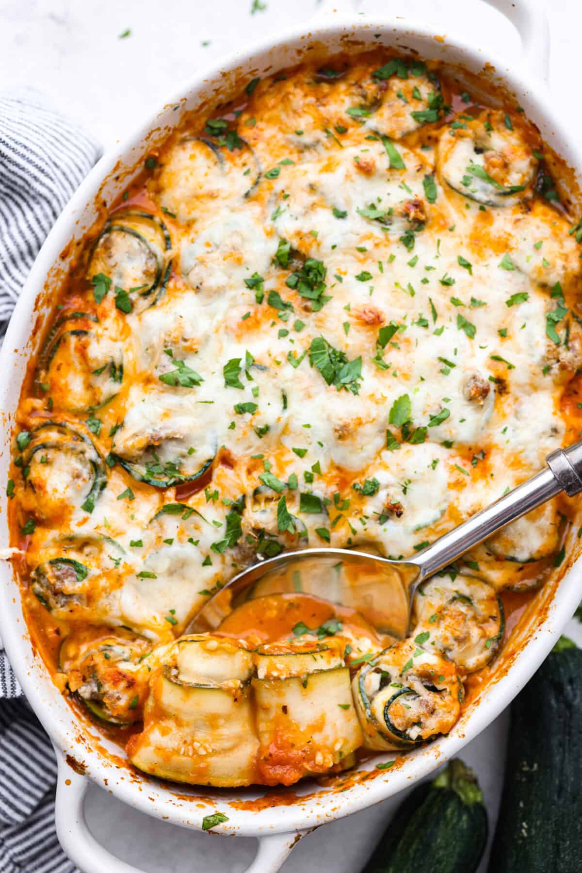 Zucchini Roll-Ups are a delicious, unique meal that the whole family will enjoy. Thin slices of zucchini are rolled around a tasty sausage ricotta mixture and layered over marinara before being topped with cheese and baked to bubbly perfection! Overhead shot of zucchini roll ups completely cooked with larger silver spoon.