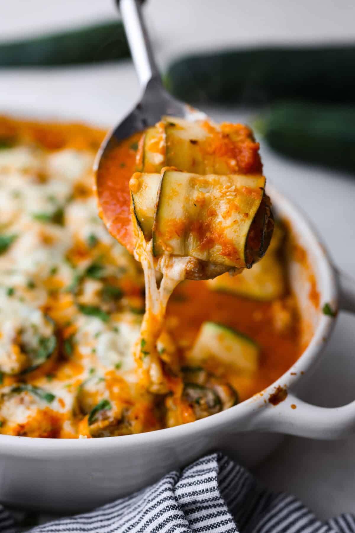 Zucchini Roll-Ups are a delicious, unique meal that the whole family will enjoy. Thin slices of zucchini are rolled around a tasty sausage ricotta mixture and layered over marinara before being topped with cheese and baked to bubbly perfection! Side shot of two zucchini rolls on a large spoon being served out of the pan.